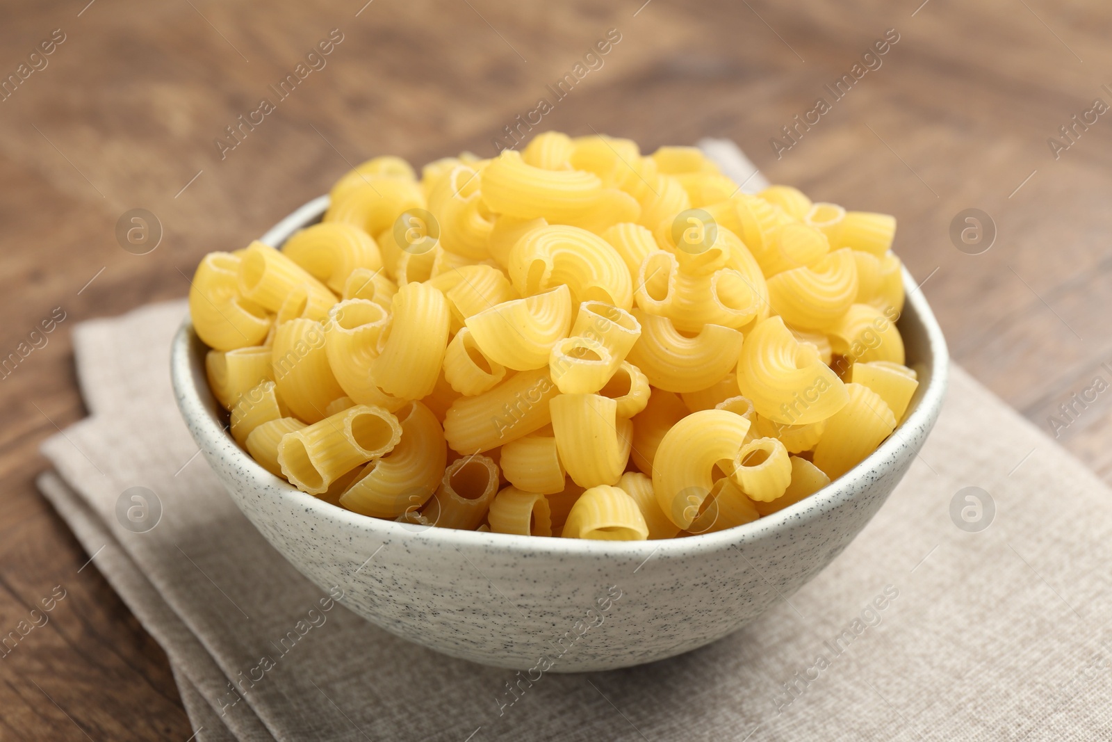 Photo of Raw horns pasta in bowl on brown table, closeup