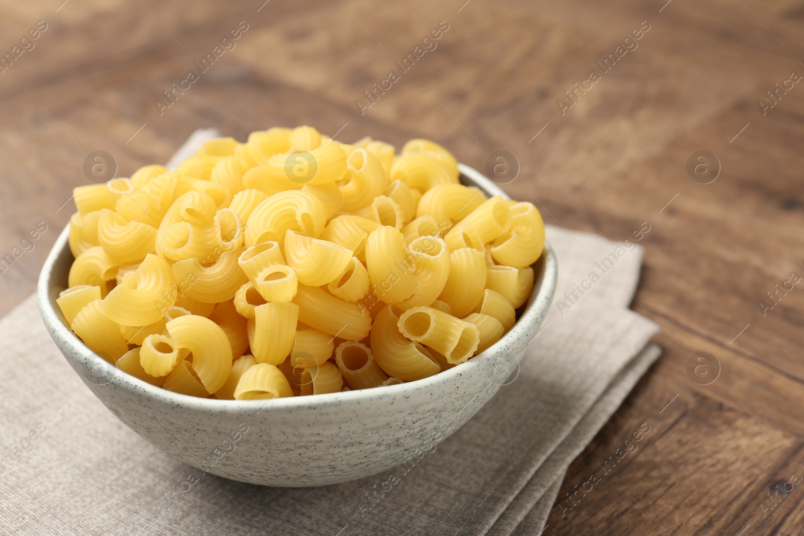 Photo of Raw horns pasta in bowl on wooden table, closeup