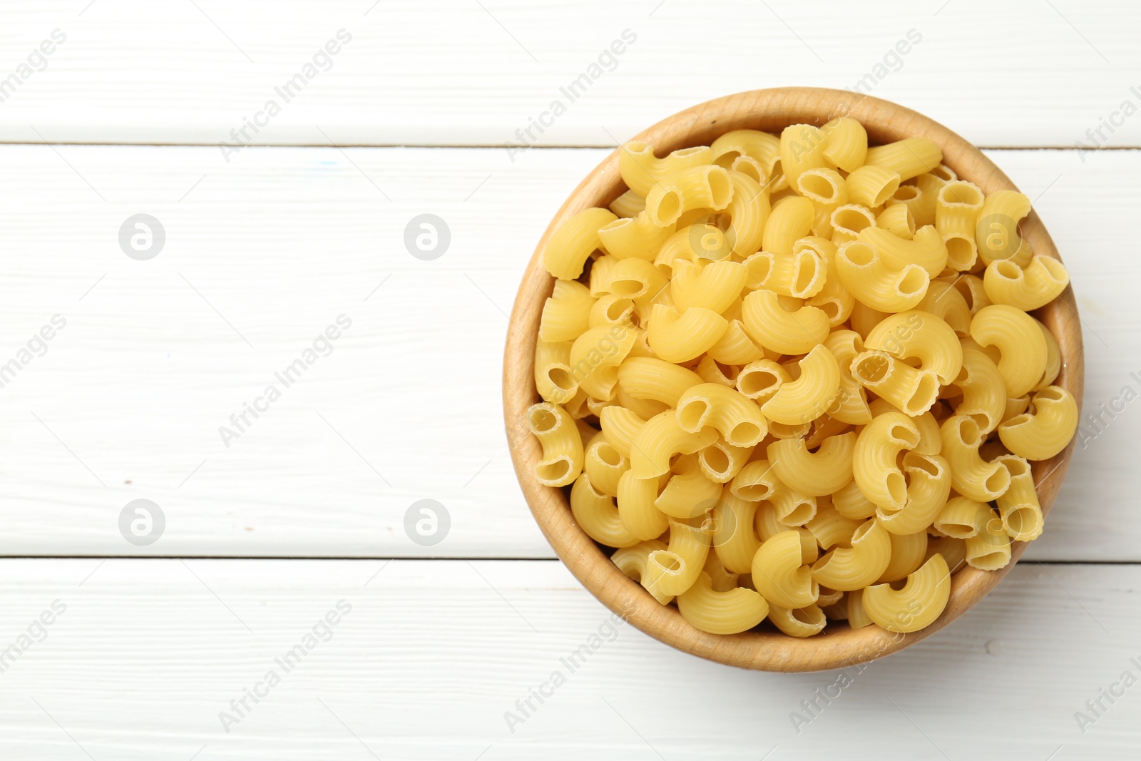 Photo of Raw horns pasta in bowl on white wooden table, top view. Space for text