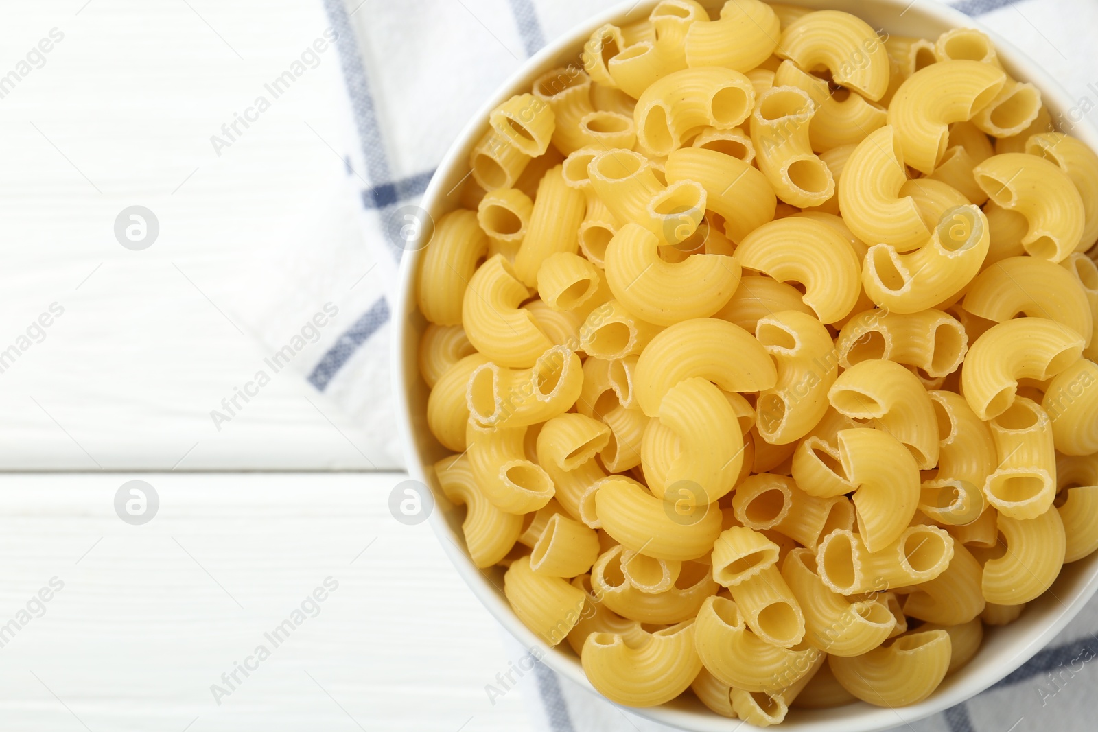 Photo of Raw horns pasta in bowl on white wooden table, top view. Space for text