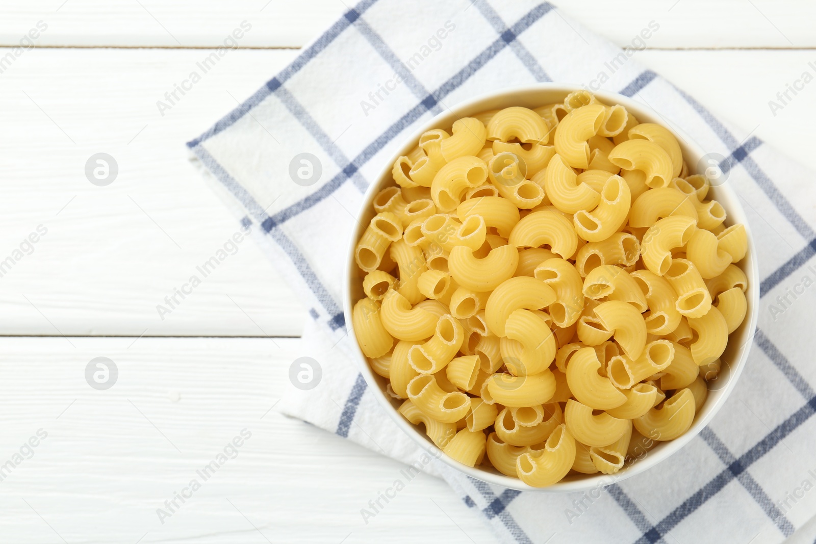 Photo of Raw horns pasta in bowl on white wooden table, top view. Space for text