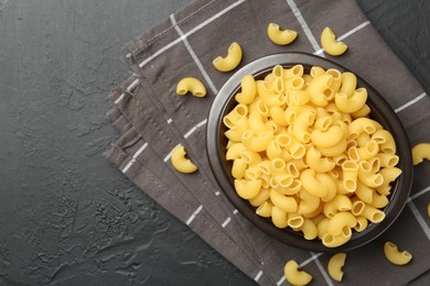 Photo of Raw horns pasta in bowl on black textured table, top view. Space for text