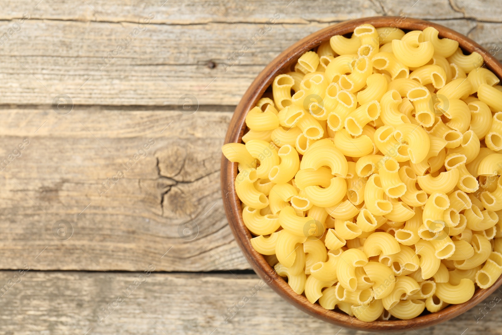 Photo of Raw horns pasta in bowl on wooden table, top view. Space for text