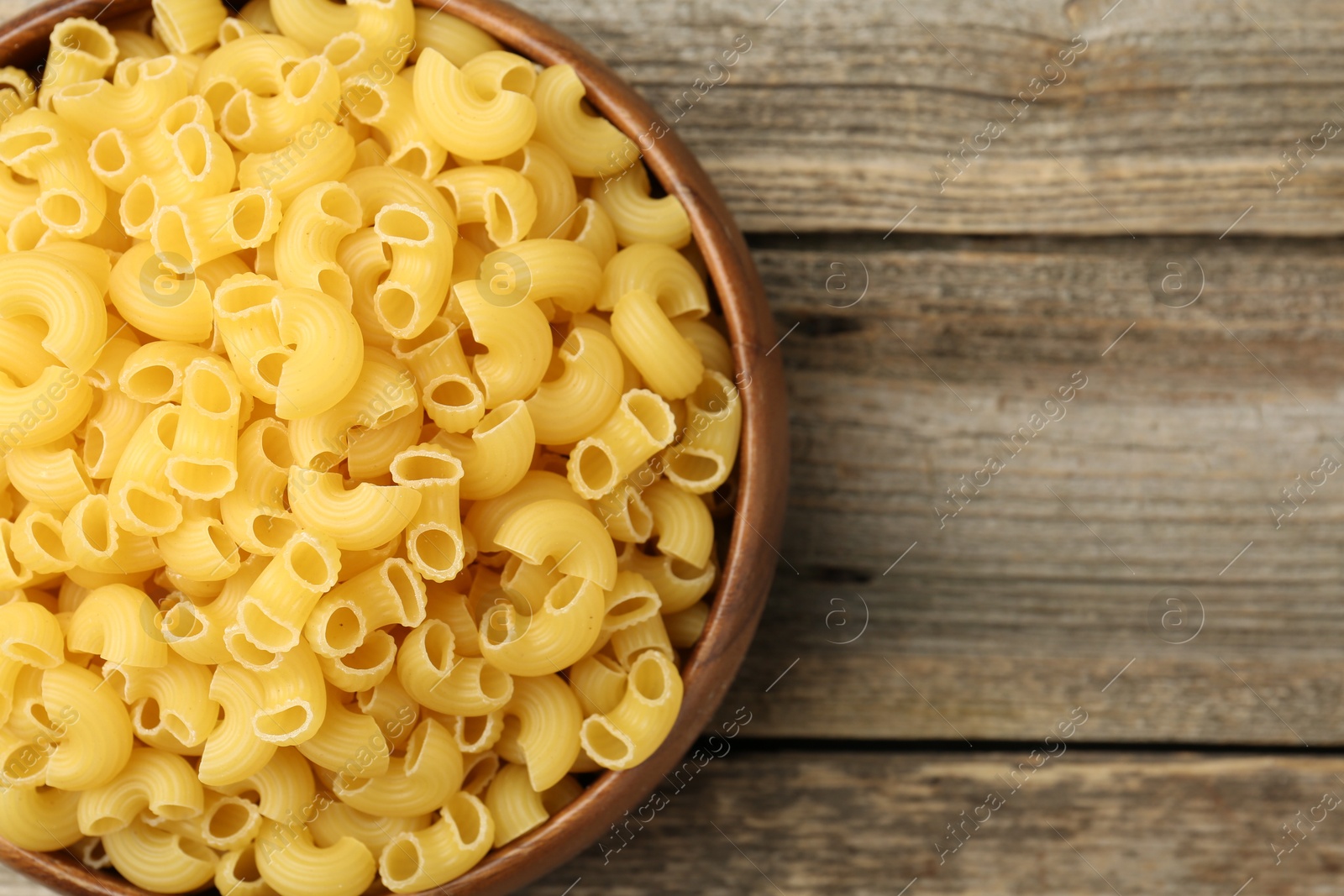 Photo of Raw horns pasta in bowl on wooden table, top view. Space for text