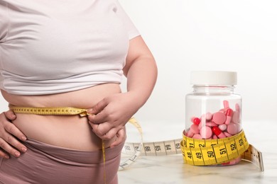 Image of Weight loss. Overweight woman measuring her waist with tape and pills on white background, closeup