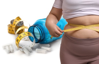 Image of Weight loss. Overweight woman measuring her waist with tape and pills on white background, closeup