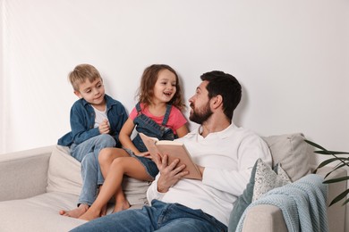 Photo of Overwhelmed father reading book to his children on sofa at home