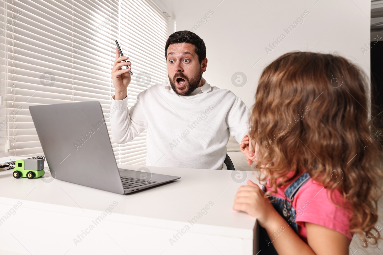 Photo of Naughty daughter disturbing her overwhelmed father at table with laptop indoors