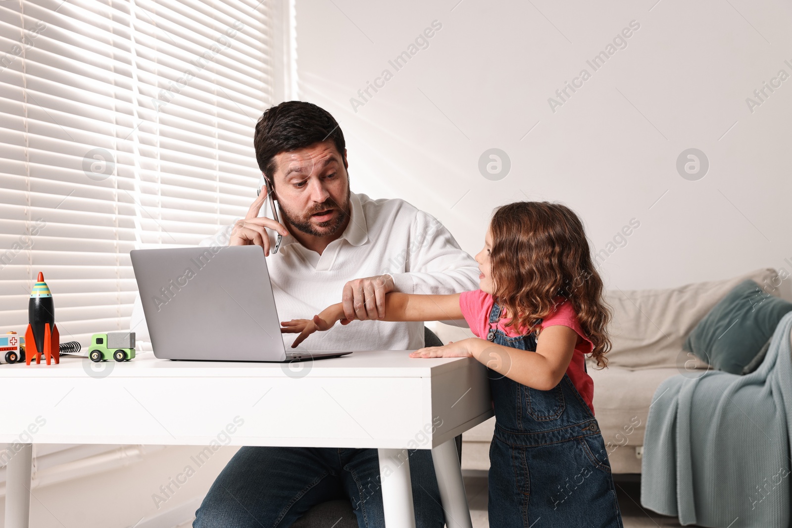 Photo of Naughty daughter disturbing her overwhelmed father while he talking on smartphone at table indoors