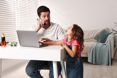 Photo of Naughty daughter disturbing her overwhelmed father while he talking on smartphone at table indoors