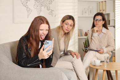 Photo of Rebellious teenage girl filming video during consultation with her mom and psychologist in office, selective focus