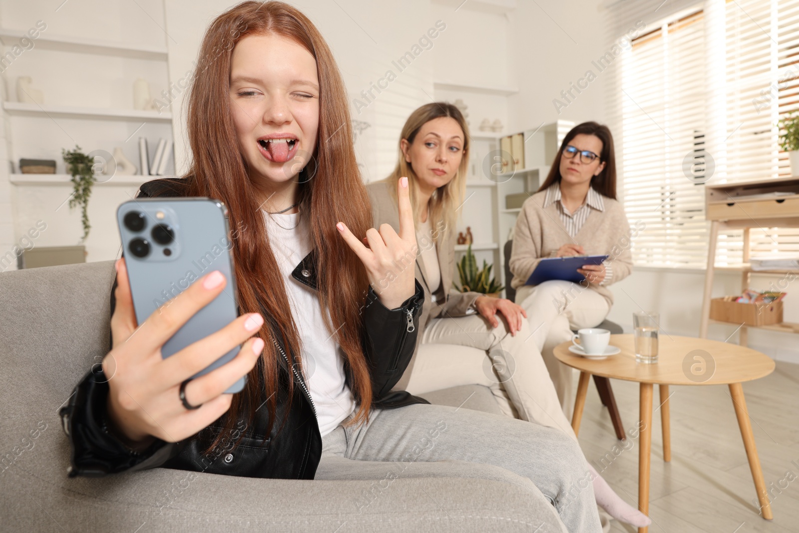 Photo of Rebellious teenage girl filming video during consultation with her mom and psychologist in office, selective focus