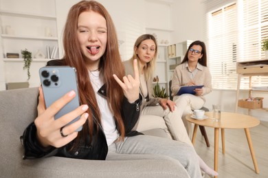 Photo of Rebellious teenage girl filming video during consultation with her mom and psychologist in office, selective focus