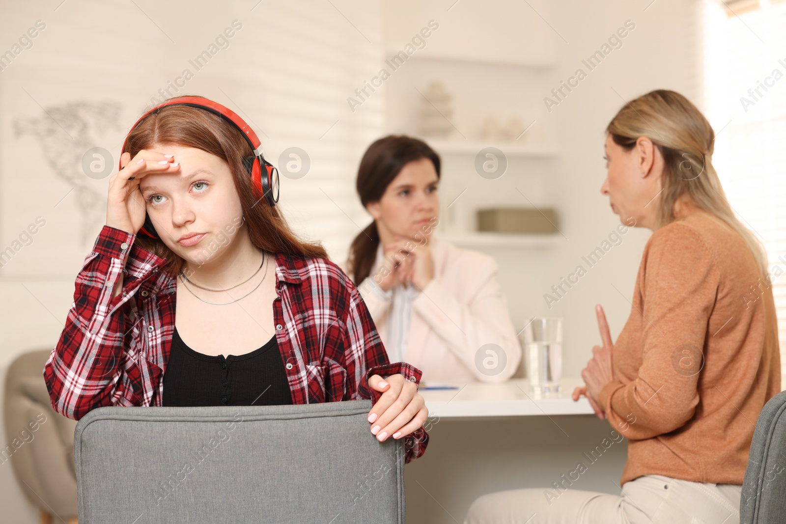 Photo of Annoyed teenage girl listening to music instead of working with psychologist in office, selective focus. Mother and daughter difficult relationship