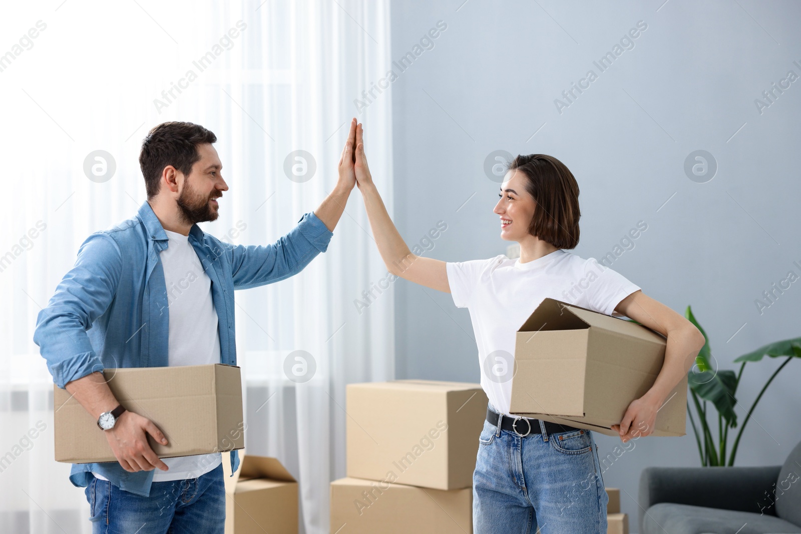 Photo of Moving day. Happy couple with cardboard boxes giving each other high five in their new home