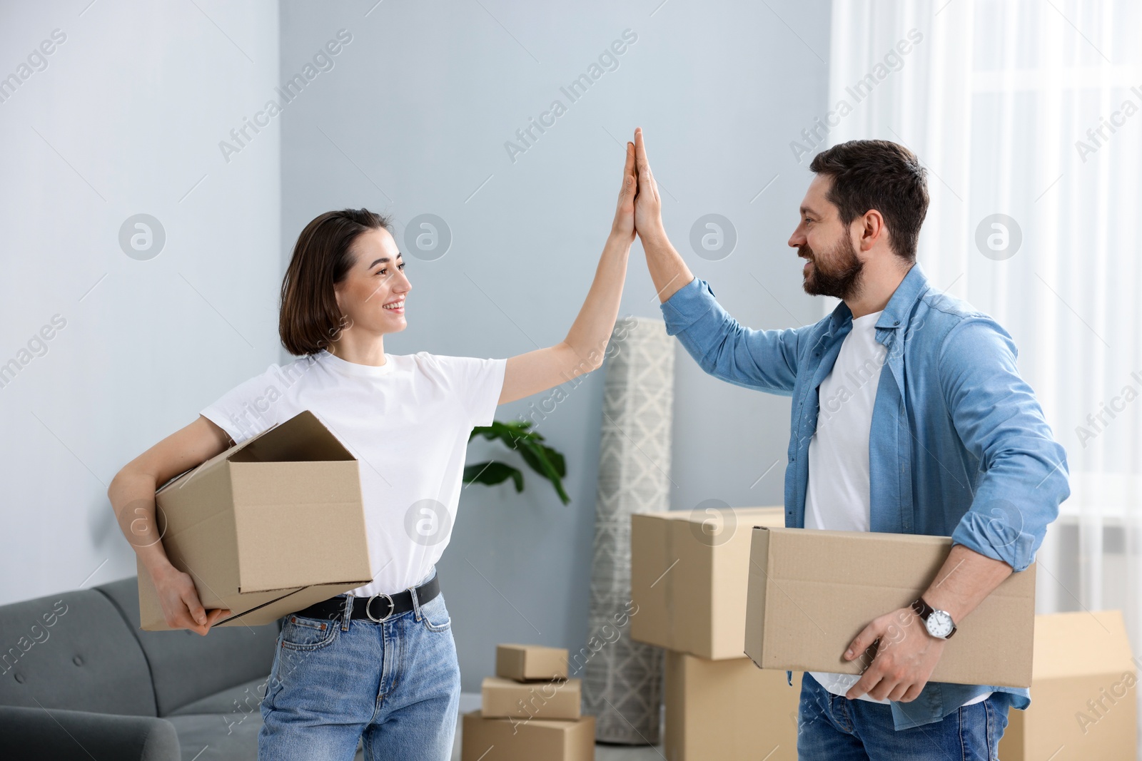 Photo of Moving day. Happy couple with cardboard boxes giving each other high five in their new home