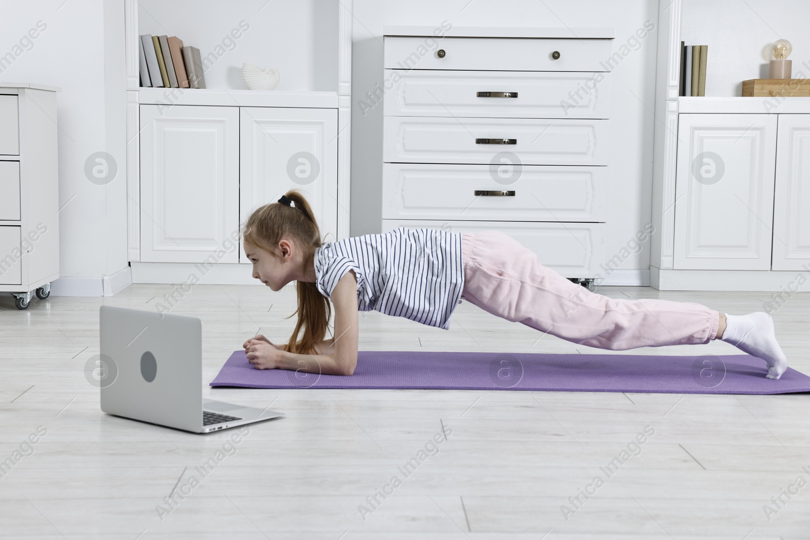 Photo of Little girl doing plank exercise near laptop at home. Morning routine