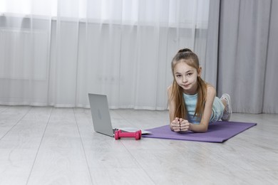 Photo of Little girl doing plank exercise near laptop at home, space for text. Morning routine