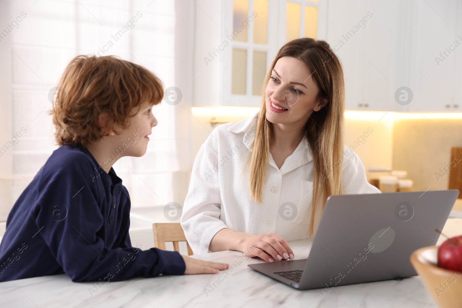 Photo of Smiling woman and her son wearing stylish pajamas with laptop at table in kitchen