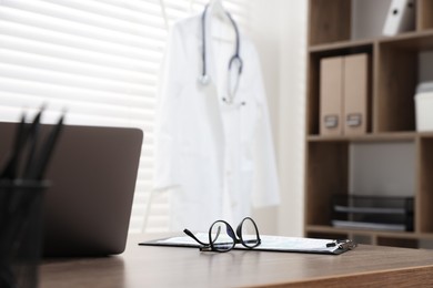 Photo of Laptop, clipboard and glasses on desk in medical office, closeup