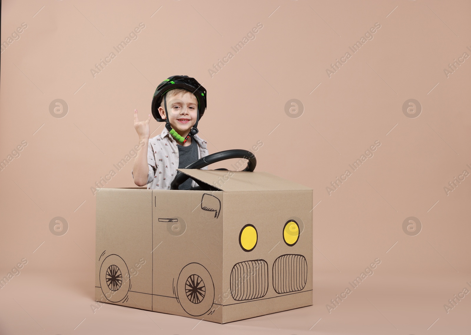 Photo of Little boy showing sign of horns while driving car made with cardboard on beige background. Space for text