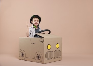 Photo of Little boy showing sign of horns while driving car made with cardboard on beige background. Space for text