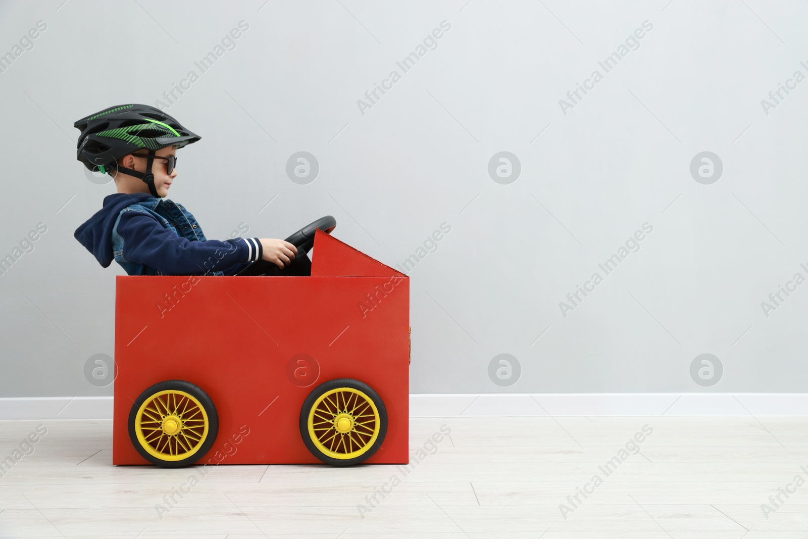 Photo of Little boy driving car made of cardboard against light wall. Space for text