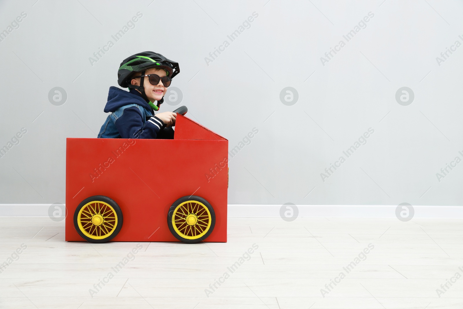 Photo of Little boy driving car made of cardboard against light wall. Space for text