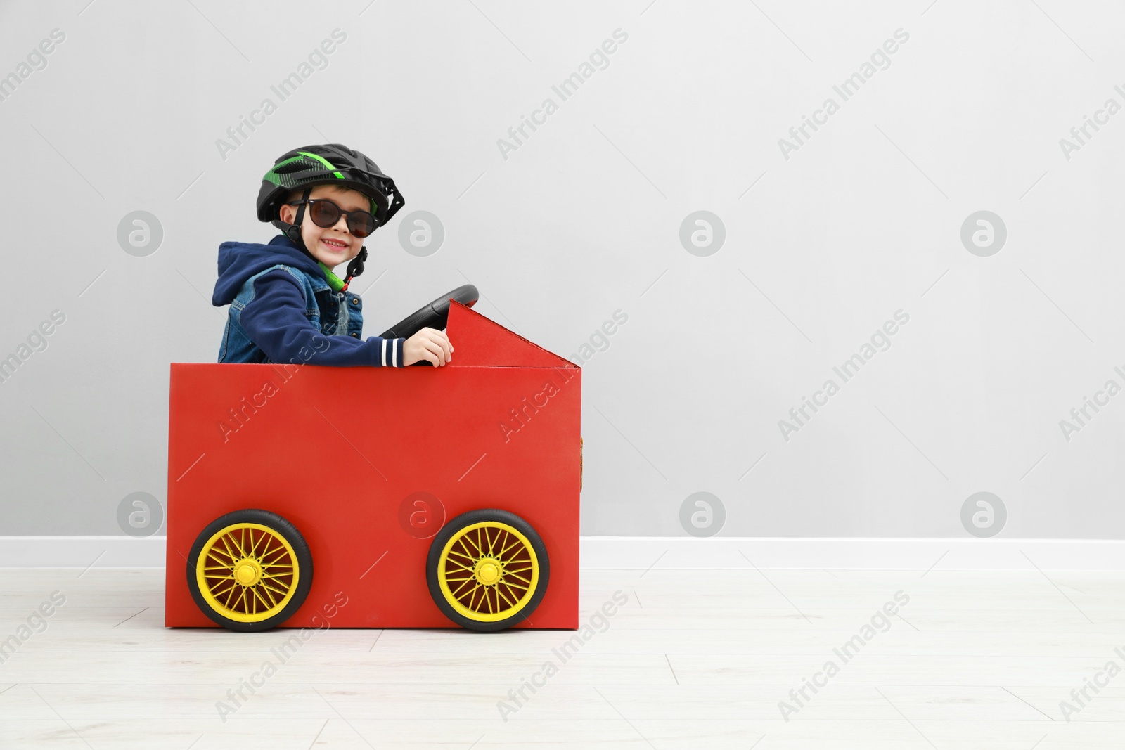 Photo of Little boy driving car made of cardboard against light wall. Space for text