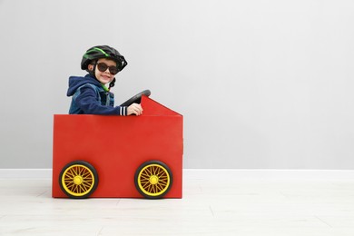 Photo of Little boy driving car made of cardboard against light wall. Space for text