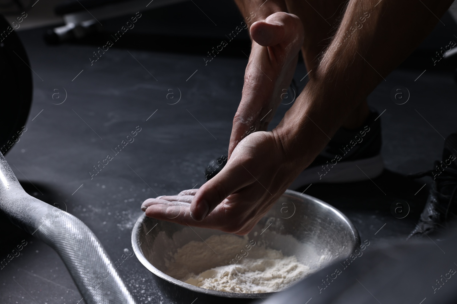 Photo of Man applying talcum powder onto his hands above bowl before training with barbell in gym, closeup