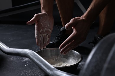 Photo of Man applying talcum powder onto his hands above bowl before training with barbell in gym, closeup