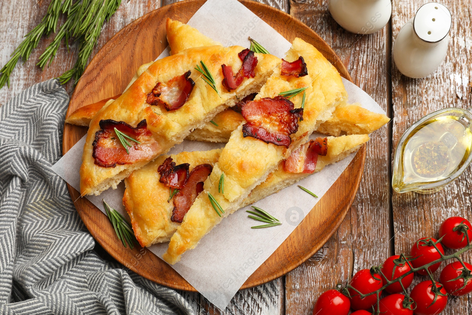 Photo of Slices of delicious focaccia bread with bacon, rosemary, oil and tomatoes on wooden table, flat lay