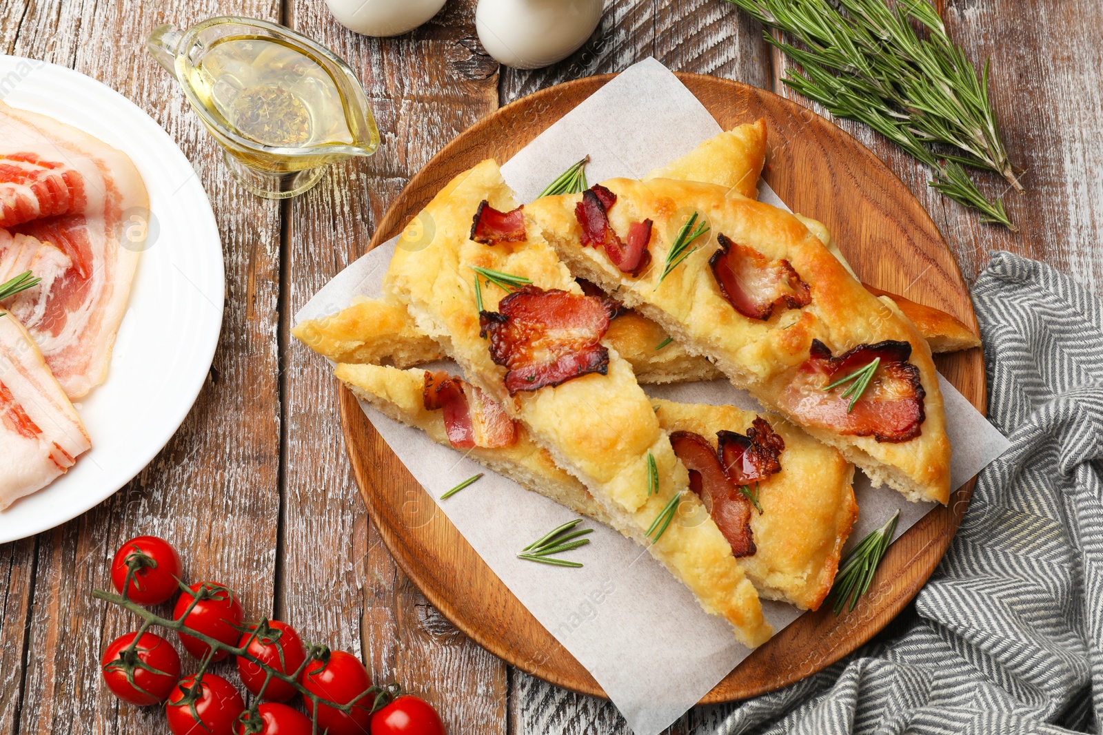 Photo of Slices of delicious focaccia bread with bacon, rosemary, oil and tomatoes on wooden table, flat lay