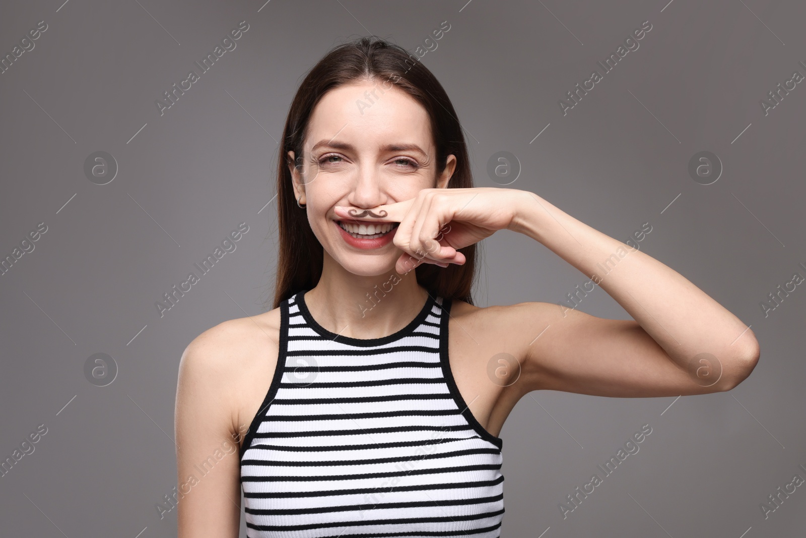 Photo of Happy woman holding finger with drawn mustache above lips on grey background