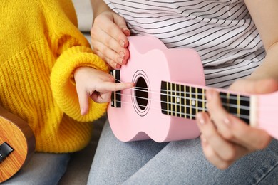 Photo of Woman teaching little girl to play ukulele at home, closeup