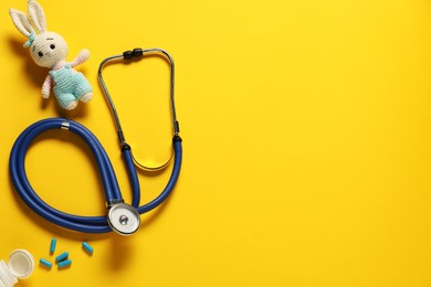 Photo of Pediatrics concept. Toy bunny with stethoscope and pills on yellow background, flat lay. Space for text