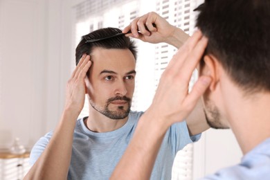 Photo of Handsome man styling his hair with comb near mirror at home