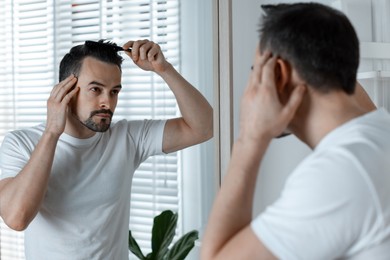 Photo of Handsome man styling his hair with comb near mirror at home