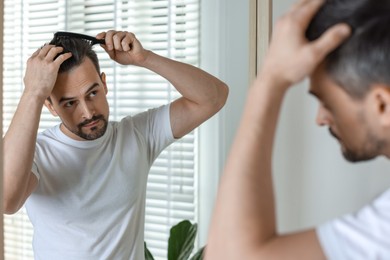 Photo of Handsome man styling his hair with comb near mirror at home