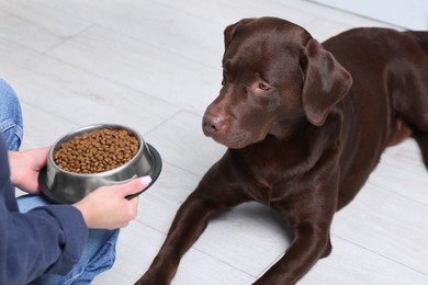 Photo of Woman giving bowl with dry pet food to her dog indoors, closeup