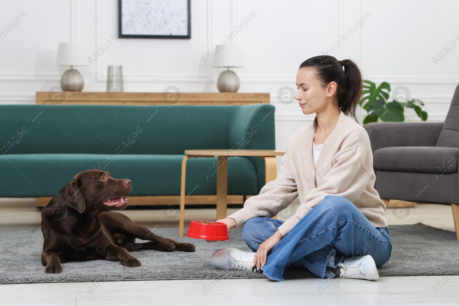 Photo of Woman giving bowl with dry pet food to her dog on floor indoors