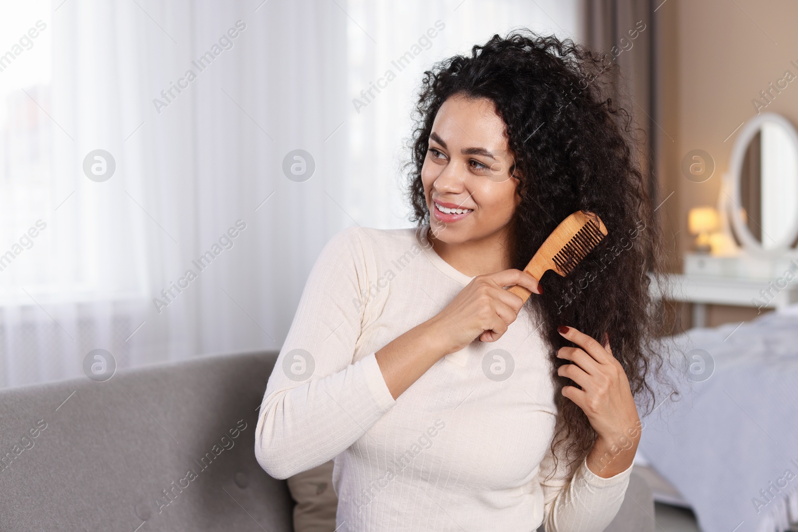 Photo of Smiling young woman brushing her curly hair with comb at home