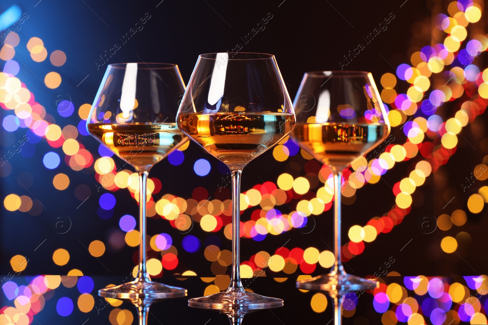 Photo of Tasty white wine in glasses on mirror surface against dark background with blurred lights, bokeh effect