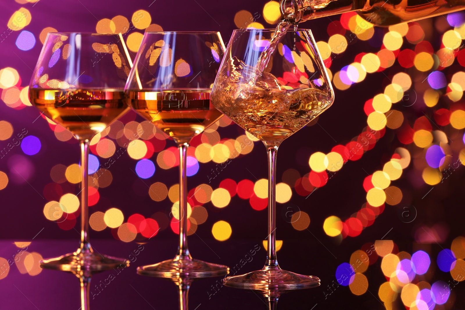 Photo of Pouring white wine into glass against purple background with blurred lights, closeup. Bokeh effect