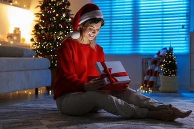 Photo of Happy woman in Santa hat with Christmas gift on floor at home