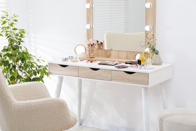 Photo of Makeup room. Dressing table with mirror, different beauty products, jewelry, armchair and houseplant indoors