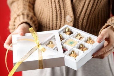 Photo of Woman holding box with tasty chocolate candies in shape of baubles on red background, closeup