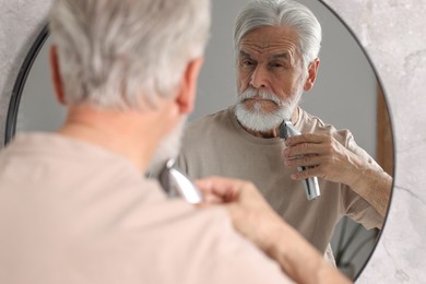 Photo of Senior man trimming beard near mirror in bathroom