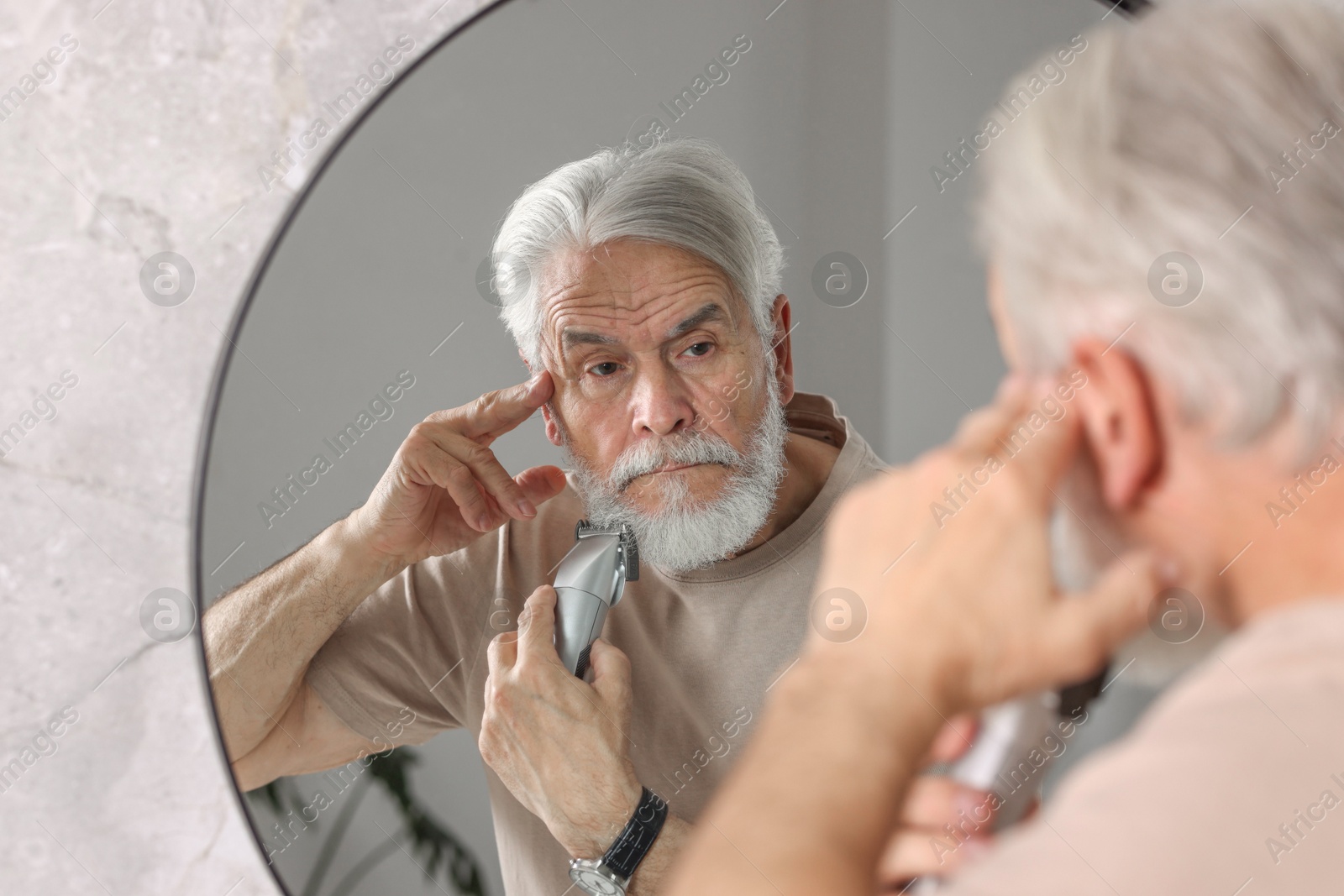 Photo of Senior man trimming beard near mirror in bathroom